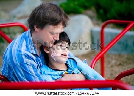 Father playing with disabled son on merry go round at playground. Child has cerebral palsy.