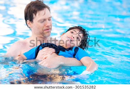 Caucasian father swimming in pool with biracial disabled son in his arms. Child has cerebral palsy.