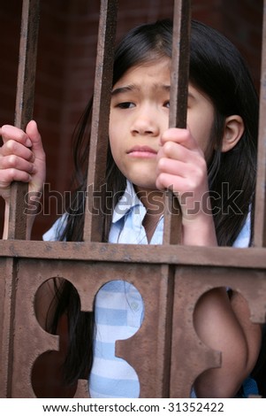 Little girl standing behind iron bars with sad expression, wither looking in or out of gate or prison