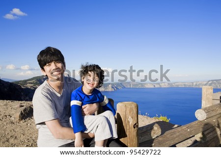 Big brother holding smiling disabled little boy with Oregon\'s famous Crater Lake in the background. Child has cerebral palsy