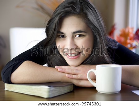 Beautiful biracial teenage girl at table with chin on arm, relaxing with a cup of coffee and her Bible or book