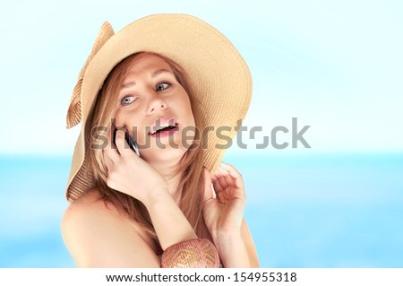Beautiful blond woman in a summer hat talking on a smart-phone, laughing on the beach