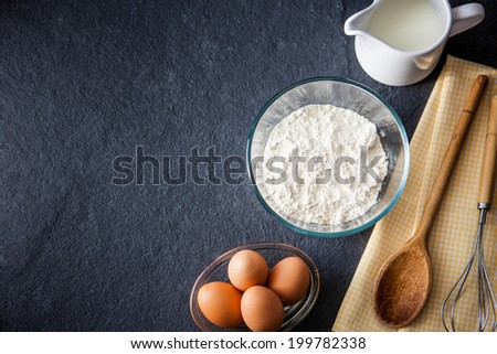 Baking ingredients - flour, milk, eggs with a whisk, wooden spoon and napkin on a slate background