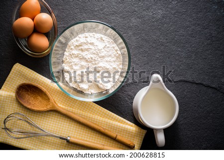 Baking ingredients - flour, milk, eggs with a whisk, wooden spoon and napkin on a slate background with copy space