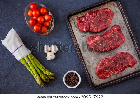 Three raw fillet steaks with green asparagus, mushrooms, cherry tomatoes with a slate background