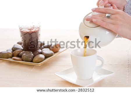 A cup of tea being poured from a white tea pot with a lit candle and stones in the background
