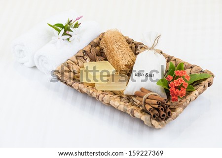 Bath salt in cotton pouch, soaps, loofah and cinnamon sticks in banana leaf basket and fluffy towels on luxurious white striped bed linen close up