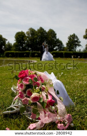 Bridal Bouquet with her shoes on meadow in a park. Bridal couple in the distance and diffuse. Focus on flowers.