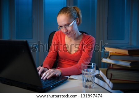 young  happy female college student typing on her notebook at late evening, chatting on social network instead of studying, funny night procrastination concept