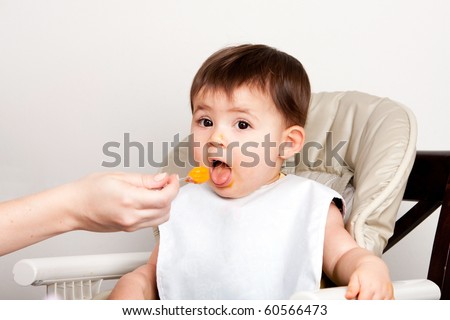 Beautiful happy baby infant boy girl with mouth open eating messy orange puree from spoon.