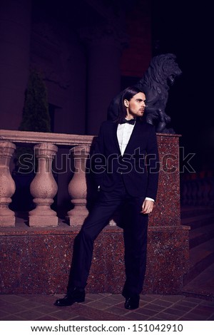 Portrait of fashion elegant long-haired young caucasian sexy muscled attractive handsome man model businessman in black costume with moustache in the street at night