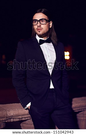 Portrait of fashion elegant long-haired young caucasian sexy muscled attractive handsome man model businessman in black costume with moustache in the street at night in glasses