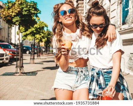 Two young beautiful smiling hipster female in trendy summer clothes. Carefree women posing outdoors.Positive models holding and drinking fresh cocktail smoothie drink in plastic cup with straw
