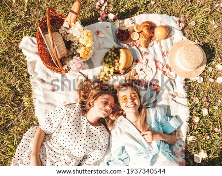 Two young beautiful smiling hipster female in trendy summer sundress and hats.Carefree women making picnic outside.Positive models sitting on plaid on grass, hugging, eating fruits and cheese.Top view