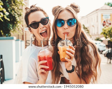 Two young beautiful smiling hipster female in trendy summer clothes. Carefree women posing outdoors.Positive models holding and drinking fresh cocktail smoothie drink in plastic cup with straw Foto d'archivio © 