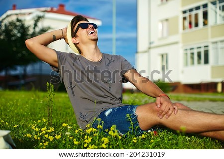 portrait of young smiling attractive modern stylish man in casual cloth in hat in sunglasses sitting in the park in green grass