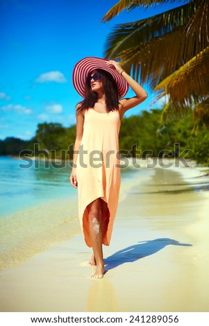 Hot beautiful woman in colorful sunhat and dress walking near beach ocean on hot summer day near palm