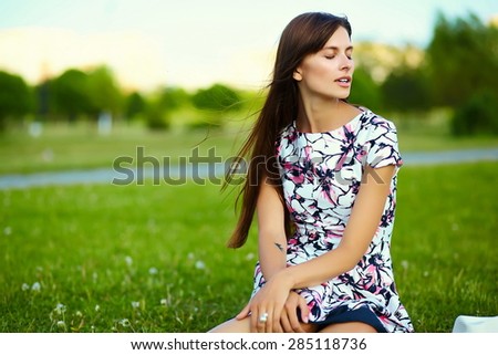 Funny  stylish sexy smiling beautiful young woman model in summer bright  hipster cloth dress with closed eyes sitting on grass in the park