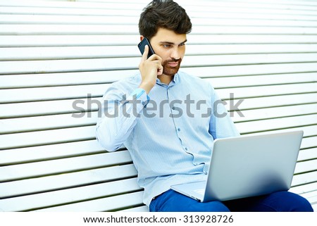 Young handsome serious businessman model sitting on the park bench using laptop in casual hipster cloth talking on mobile phone