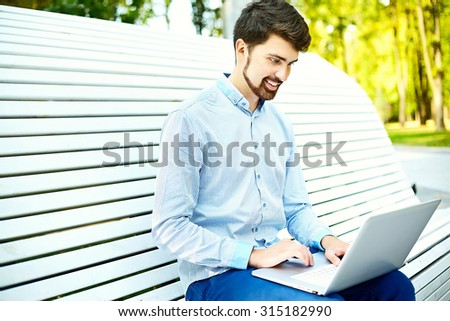 Young handsome smiling businessman model sitting on the park bench using laptop in casual hipster cloth