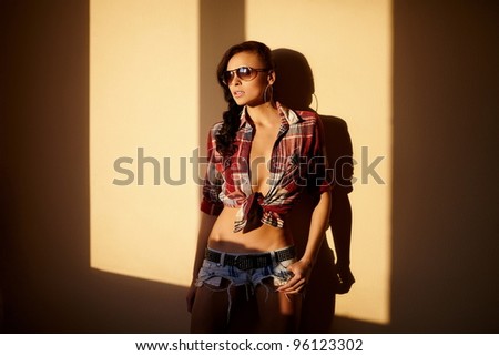 portrait of beautiful fashion sexy brunette girl in jeans and colorful shirt in glasses behind sunset indoors
