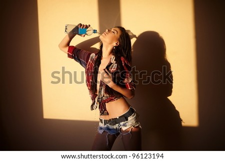 portrait of beautiful fashion sexy brunette girl in jeans and colorful shirt  behind sunset with bottle water  indoors