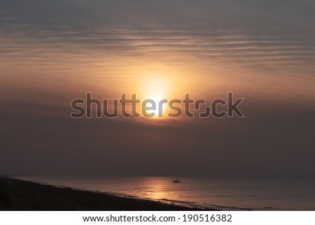A picture of the beach near sunset with people walking on the sand and in the water and a small boat on the water in the background
