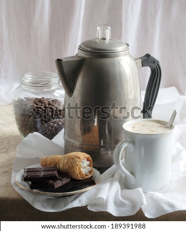 Still-life with a coffee pot and fragrant coffee with cream in a white mug both dark chocolate and an appetizing cake in the form of a tubule from flaky pastry with an albuminous cream