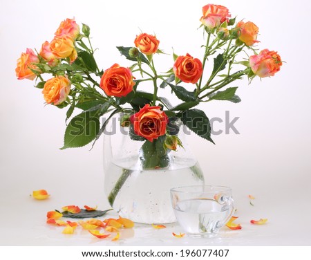 Still-life with a transparent cup and a beautiful bouquet from bright orange roses in a transparent jug