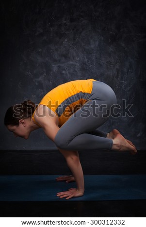 Beautiful sporty fit yogini woman practices yoga asana kakasana - crow pose on dark background