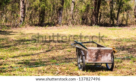 A garden cart and tools with gloves hanging off the side as it sits in a lawn ready to go to work.