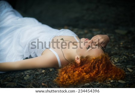 Beautiful redhead woman at the rocky beach in a white dress laid down on the gravel.