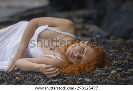 Beautiful redhead woman at the rocky beach in a white dress laid down on the gravel.