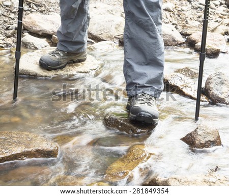 Male Hiking Over the Wild Creek Water Stones with Mountain Boots and Trekking Poles