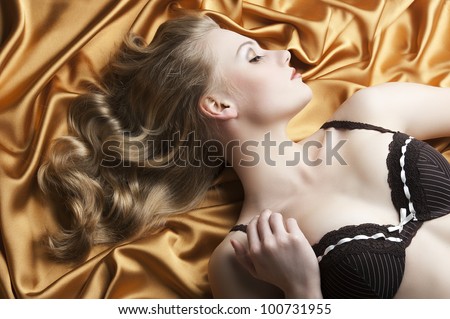 close up portrait of blond beautiful girl with well done hair style laying down on golden shining material, she is turned in profile at left and she looks down, her right hand is near the right
