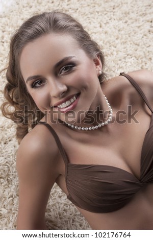 portrait of a young sexy girl in brown bra with a pearl necklace laying on a white modern carpet and smiling