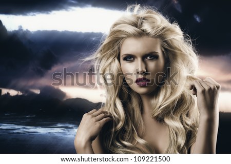 portrait of young sexy girl with blode wavy hairstyle and and flying hair from wind, she is in front of the camera and looks in to the lens, her right hand is near the right shoulder