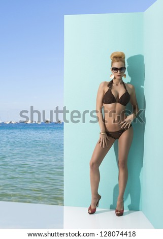 blonde sexy woman in bikini with blonde hair, heels and sunglasses posing near wall. Blue sky, sea and some boats on the background