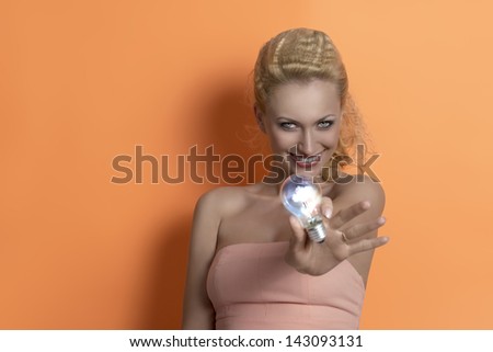 pretty blonde woman with pink dress and pretty hair-style showing light bulb and smiling.  On orange background