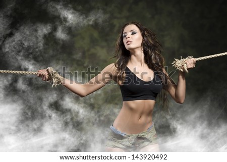 sensual aggressive girl with fitness body and tied arms by rope wearing dark top and shorts , with smoke and military background