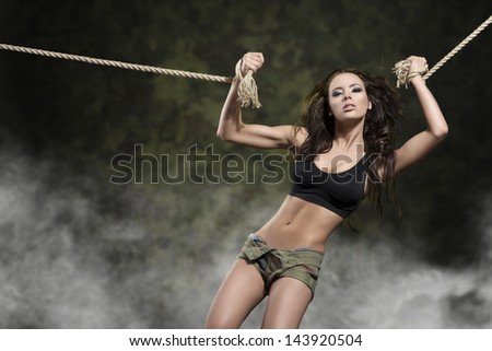 sexy woman with perfect body and brown flying hair is tied by rope and wearing black top and shorts on military background