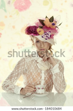 blonde girl with floral hat and vintage shirt behind the table with cup of tea, she looks in to the lens and her left hand is under the chin