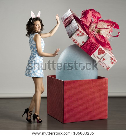 very cute young woman in spring ligh blue dress, bunny ears near big red box with easter egg inside