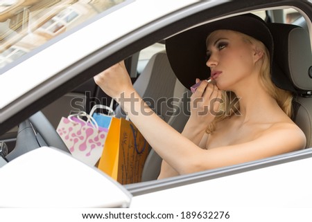 beauty woman sitting in car , with hat and shopping bags near, in act to make up her lips.