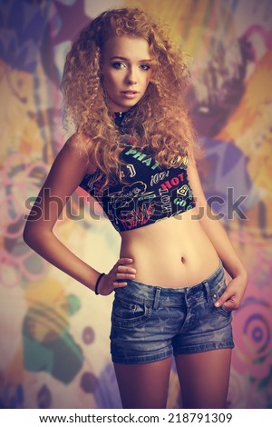 fashion portrait of very pretty teenager girl with curly hair and casual clothes, posing with trendy top and denim shorts, looking in camera with innocent expression