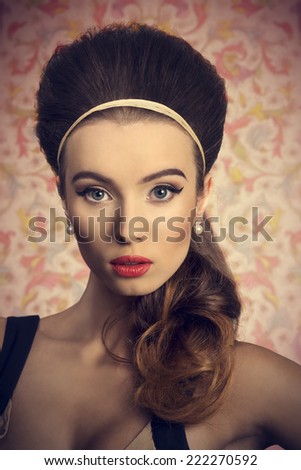 Short portrait of young, beautiful woman in fitted cream and black dress is looking down. She has got big blue eyes, brown big hairbun and she is wearing hairband and pearls on her ears.