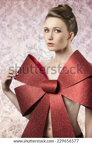 sensual woman with elegant blonde hair-style posing in christmas shoot with big red glitter bow covering her naked breast, looking in camera