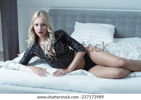 sexy young woman with long blonde hair posing on bed with black panties and leather jacket, erotic neckline looking in camera