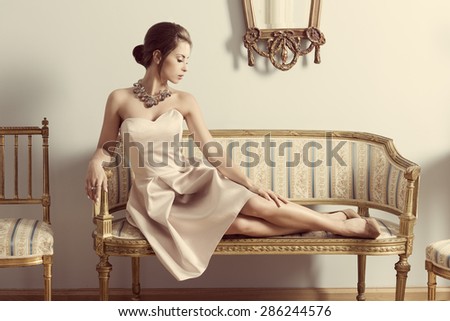 interior portrait of brunette elegant girl lying on retro sofa in aristocratic room. Wearing pink dress, precious jewellery and classic hair-style. Luxury atmosphere