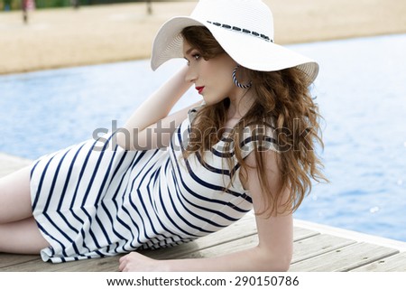 fashion woman with trendy striped dress and hat lying with romantic expression on wood jetty on blue sea water. Fashion summer portrait of cool girl in vacation